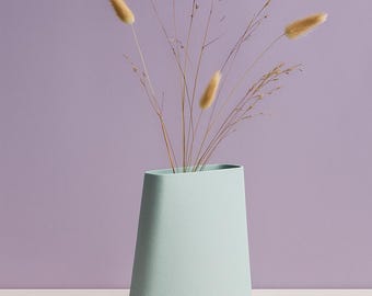 May include: A light blue vase with a unique, asymmetrical shape. The vase holds dried ornamental grasses with fluffy, tan seed heads. The background is a solid lavender color, and the vase sits on a white surface. A modern, minimalist design.