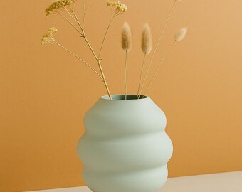 May include: A light green, ceramic vase with a unique, rounded design. The vase holds dried flowers and grasses. The background is a solid, warm orange color, and the vase sits on a neutral surface. The vase is approximately 10 inches tall.