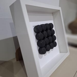 Può includere: Una cornice bianca con un'opera d'arte astratta in bianco e nero al suo interno. L'opera d'arte è composta da piccoli oggetti rotondi neri disposti in una griglia.