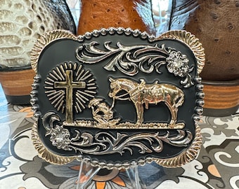 Puede incluir: Una hebilla de cinturón negra y plateada con un diseño dorado que presenta una cruz, un caballo y un jinete. La hebilla tiene un fondo negro y un borde plateado.