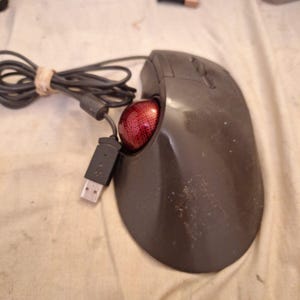 May include: A dark gray computer trackball mouse featuring a prominent red ball with a textured grid pattern. The device includes a black integrated cable with a standard USB connector, designed for wired connectivity to a computer system.