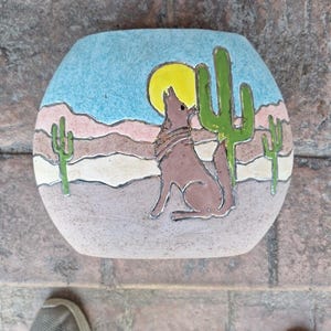 Puede incluir: Un jarrón de cerámica con una escena del desierto pintada en él. La escena incluye un coyote aullando a la luna, un cactus y una puesta de sol. El jarrón es de color azul claro con acentos marrones, rosados y verdes.