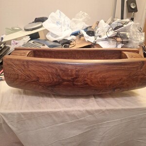 May include: A long, rectangular wooden bowl with a deep, curved interior and a rich, dark brown wood grain. The exterior features subtle, carved leaf-like patterns on both sides, with a smooth, polished finish and a sturdy, solid construction.