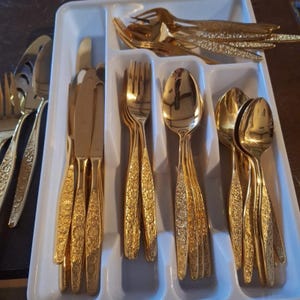 May include: A set of gold-colored flatware, including forks, knives, and spoons, arranged in a white tray. The flatware has a decorative floral pattern. The image shows a variety of utensils, including serving forks and spoons.