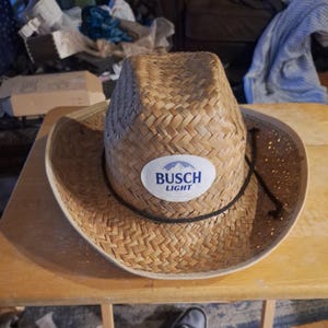 Puede incluir: Un sombrero de vaquero de paja tejida color canela con un parche ovalado blanco en la parte delantera que muestra el texto azul Busch Light y un gráfico de montaña. El sombrero incluye un cordón negro fino alrededor de la base y ala ancha.