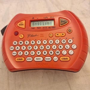 Może przedstawiać: Pomarańczowa drukarka etykiet Brother P-touch z klawiaturą QWERTY i ekranem LCD wyświetlającym "English". Funkcje obejmują przyciski drukowania, czasu/daty i czyszczenia. Urządzenie ma kompaktową konstrukcję.