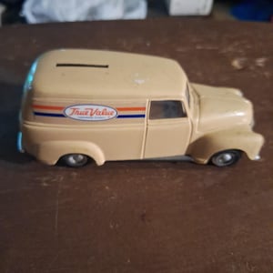 May include: A beige toy delivery van coin bank featuring the True Value logo on the side panel. The van has a horizontal slot on the roof for inserting coins, a rounded body design, and small black wheels. The logo includes orange and blue stripes.