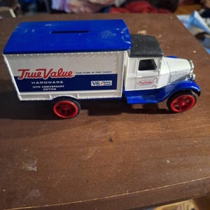 May include: A die-cast metal toy truck piggy bank featuring a blue roof with a coin slot, white cargo box, and white cab. The side displays True Value Hardware 10th Anniversary Edition branding with red wheels and a black roof on the cab section.