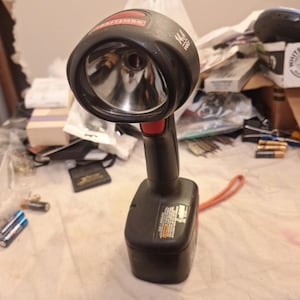 May include: A black and red Craftsman 14.4V flashlight. The flashlight has a large, round head with a reflective interior and a red accent on the handle. A red lanyard is attached to the base.