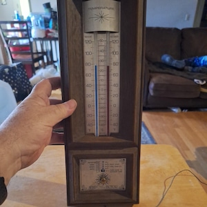 May include: A rectangular wall-mounted thermometer with a dark wood frame. The silver face displays indoor and outdoor temperatures in Fahrenheit and Celsius. Below the thermometer is a wind chill factor chart with a small circular dial.