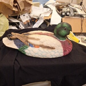 Puede incluir: Figura de pato de cerámica con cabeza verde y pico amarillo. El cuerpo presenta tonos blancos, marrones, azules y rojos. El pato está tumbado sobre una superficie negra, con detalles de plumas.