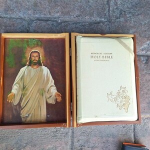 Puede incluir: Una caja de madera cerrada con una imagen de Jesucristo en el lado izquierdo y un libro blanco con el texto "Memorial Edition Holy Bible Concordance" en el lado derecho.
