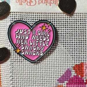 May include: A pink heart-shaped enamel pin with the text "JUST A GIRL WHO NEEDS HER LITTLE SNACKY SNACKS" in white. The pin has a black outline and blue border, with gold star accents. The pin is on a white mesh surface.