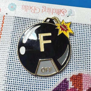 以下が含まれることがあります： 金色の縁取りが付いた黒いエナメル爆弾型のピン。金色の「F」と「OFF」の文字が灰色のパネルに表示されています。赤いアクセントが付いた黄色のスターバーストが上部に付いています。ピンは白いメッシュの背景にあります。