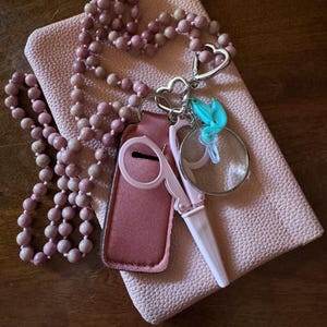 May include: A collection of pink accessories including a beaded necklace, a small pink pouch, and a pair of pink scissors with a matching case. A silver keychain with a heart design holds a small magnifying glass and a turquoise bird charm.