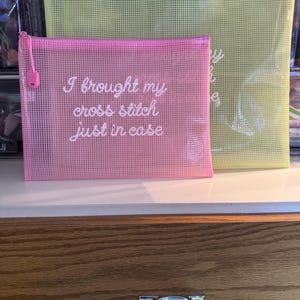 May include: Two mesh zipper pouches, one pink and one yellow, are displayed. The pink pouch has the text "I brought my cross stitch just in case" in white script. The yellow pouch is larger and has faint text. Both have a zipper.