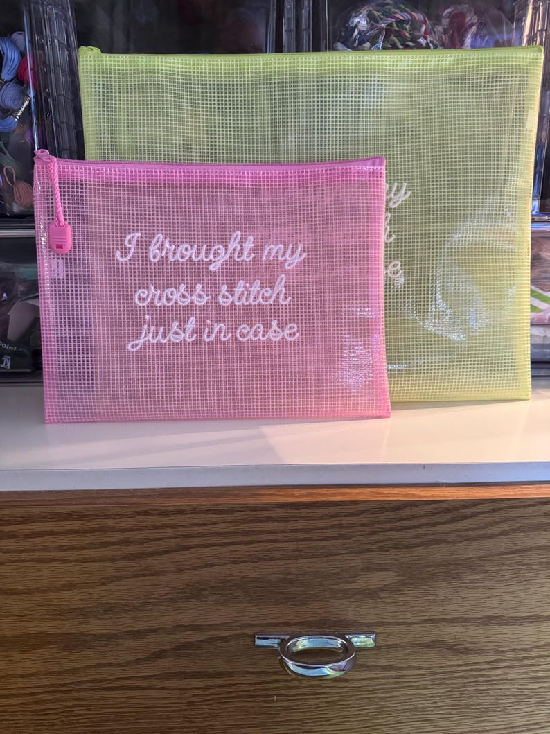 May include: Two mesh zipper pouches, one pink and one yellow, are displayed. The pink pouch has the text "I brought my cross stitch just in case" in white script. The yellow pouch is larger and has faint text. Both have a zipper.