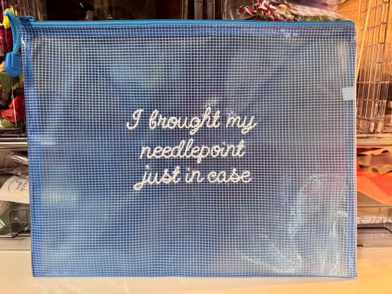 May include: A blue zippered pouch with a grid pattern and the embroidered text "I brought my needlepoint just in case." The pouch has a blue zipper and pull tab. Ideal for storing craft supplies.