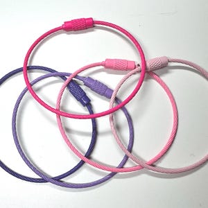 May include: Five colourful keyrings in shades of pink and purple. Each keyring has a circular wire design with a matching clasp. The keyrings are arranged in an overlapping pattern against a white background.