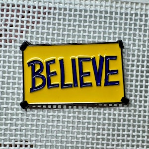 May include: A rectangular pin with the word "BELIEVE" in blue letters on a yellow background. The pin has a black border and is set against a white mesh background.