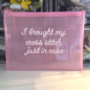 May include: A pink, mesh zipper pouch with the text "I brought my cross stitch just in case" in white script. The pouch is rectangular and appears to be made of a semi-transparent material. The background shows spools of thread.