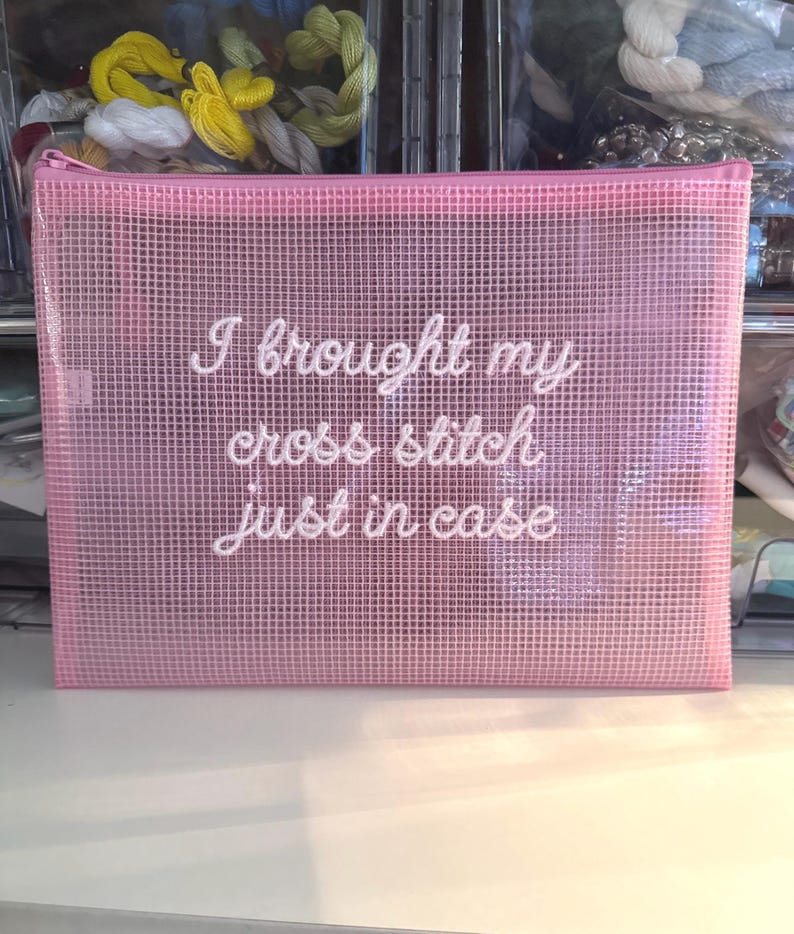 May include: A pink, mesh zipper pouch with the text "I brought my cross stitch just in case" in white script. The pouch is rectangular and appears to be made of a semi-transparent material. The background shows spools of thread.