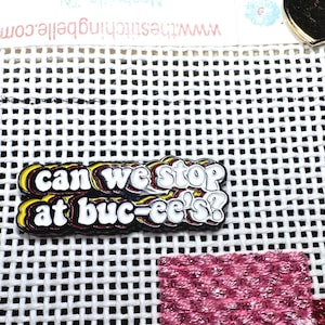 May include: A colorful enamel pin with the text "Can we stop at Buc-ee's?" in a layered, wavy font. The pin is black, white, yellow, and red. The pin is on a white grid fabric with a pink and red stitched design.