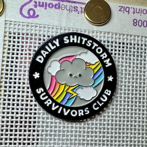 May include: Black enamel pin featuring a white cloud with a sad face and a lightning bolt, a rainbow, and the text "Daily Shitstorm Survivors Club".