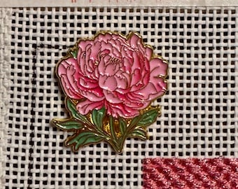 Op de afbeelding: Een goudkleurige emaille speld in de vorm van een bloeiende roze pioenroos met groene bladeren. De speld is bevestigd op een wit naaldwerkcanvas met een rasterpatroon en de tekst www.thestitchingbelle.com en Nashville, TN.