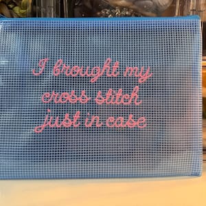 May include: A blue cross stitch project bag with a grid pattern and a zipper closure. The bag has the phrase "I brought my cross stitch just in case" embroidered in pink cursive lettering.