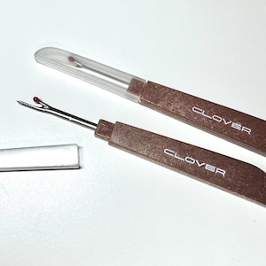 May include: Two brown seam rippers with the word "CLOVER" printed in white. One ripper has a clear plastic cap. The seam rippers are used for removing stitches. The tools are on a white surface.