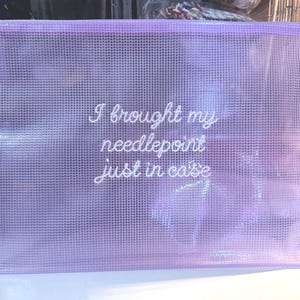 May include: A lavender mesh zippered pouch with the text "I brought my needlepoint just in case" in white script. The pouch is rectangular and appears to be made of a translucent material, with a silver zipper.