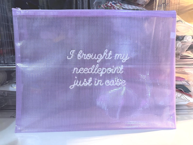 May include: A lavender mesh zippered pouch with the text "I brought my needlepoint just in case" in white script. The pouch is rectangular and appears to be made of a translucent material, with a silver zipper.