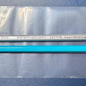 May include: A clear plastic bag contains a purple pencil sharpener, a silver pencil with the text "PRISMACOLOR VERITHIN Argent Métallique 753 Metallic Silver", and a turquoise pencil with a black end. The pencils are for drawing and sketching.
