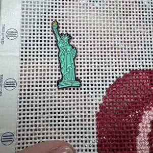 May include: A Statue of Liberty pin with a gold torch and a green body. The pin is on a white mesh canvas with the words "THREE STITCHES" printed on the side.