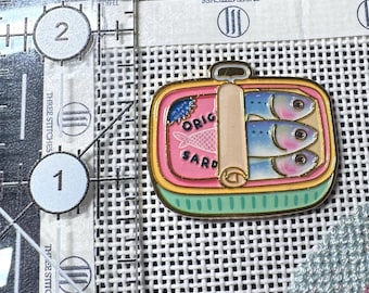 May include: A rectangular enamel pin shaped like an open tin of sardines with a pink interior, a light green base, and three blue fish. A beige tab sits across the centre. The pin is placed next to a clear ruler showing a width of 3.2 centimetres.