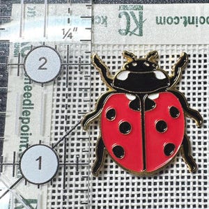 May include: A ladybug enamel pin with a red body, black spots, and a black head with white accents. The pin is positioned next to a clear ruler showing a measurement of approximately 1.25 inches in height and width for the insect figure.