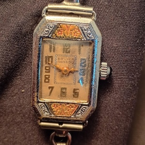 May include: Vintage rectangular wristwatch with a silver-tone case and a gold-tone face. The watch face features black numerals, gold accents, and the text "BRUNER MASTER BILT SWISS". The watch has a silver-tone band.