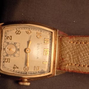Puede incluir: Reloj de pulsera Bulova vintage con una caja rectangular dorada y una esfera color crema. El reloj presenta agujas e indicadores de hora dorados, un pequeño dial de segundos y una correa de cuero marrón claro. La esfera muestra la marca Bulova.