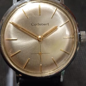 May include: A vintage Cortébert wristwatch with a gold-toned face and hands, and gold-colored hour markers. The watch has a silver-colored metal case and a black leather band. The watch face displays the brand name "Cortébert".