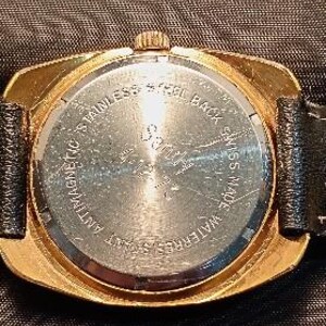 May include: Close-up of the back of a gold-toned wristwatch with a black leather band. The watch face is round with the text "STAINLESS STEEL BACK", "ANTIMAGNETIC", "WATERRESISTANT", and "SWISS MADE".