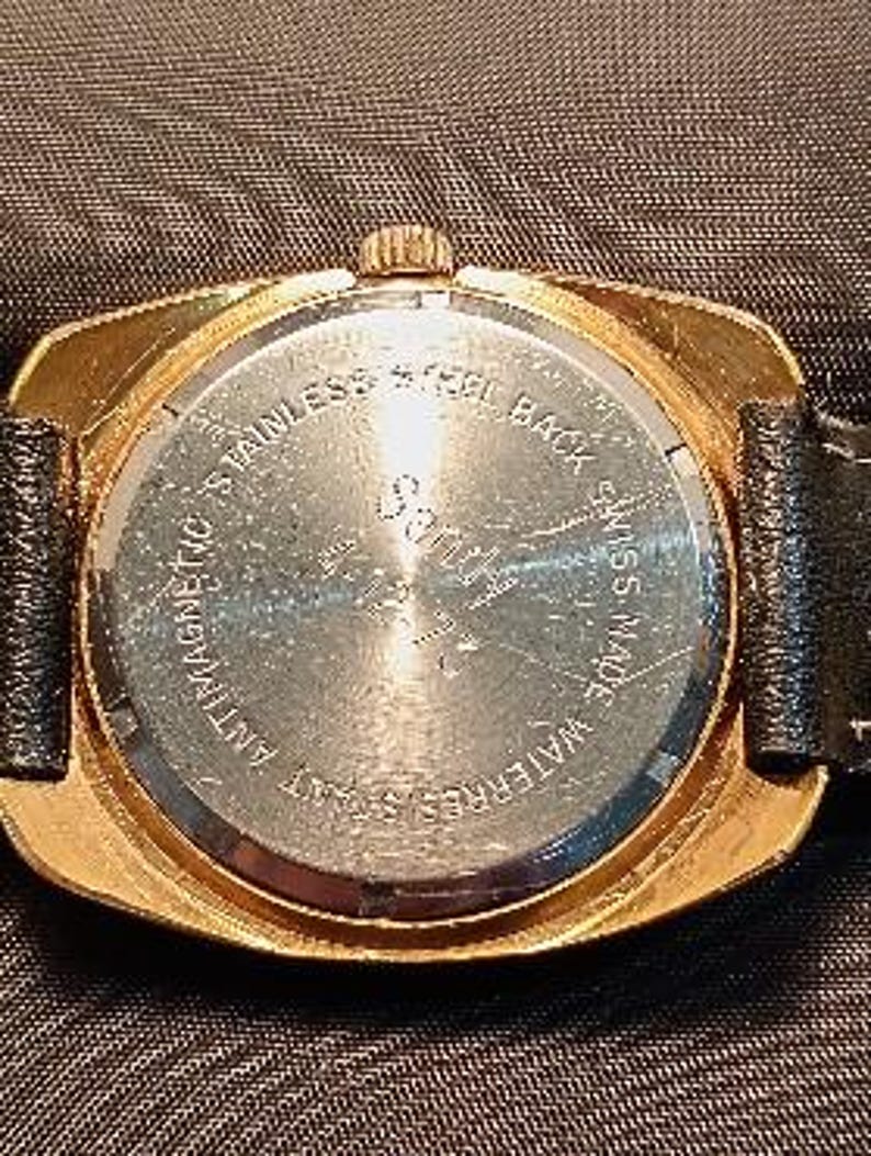 May include: Close-up of the back of a gold-toned wristwatch with a black leather band. The watch face is round with the text "STAINLESS STEEL BACK", "ANTIMAGNETIC", "WATERRESISTANT", and "SWISS MADE".