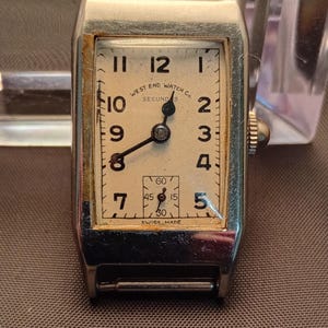 Op de afbeelding: Vintage rechthoekig polshorloge met een crèmekleurige wijzerplaat. Het horloge heeft zwarte cijfers, wijzers en de tekst "WEST END WATCH CO. SECUNDS SWISS MADE". Het heeft een zilverkleurige metalen kast en een kleine secondenwijzerplaat.