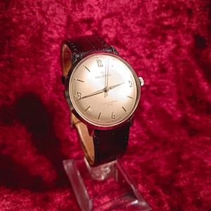 May include: A vintage Helbros Invincible watch with a silver-tone face, black leather band, and gold-tone hands and markers. The watch is displayed on a clear stand against a red background. The watch face has the number 12, 3, 6, and 9.