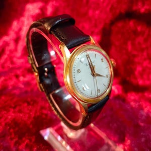 May include: A vintage gold-tone wristwatch with a white face and black leather band. The watch features gold hands and hour markers, with the numbers 2, 6, and 10 clearly visible. The watch is displayed on a clear stand against a red background.
