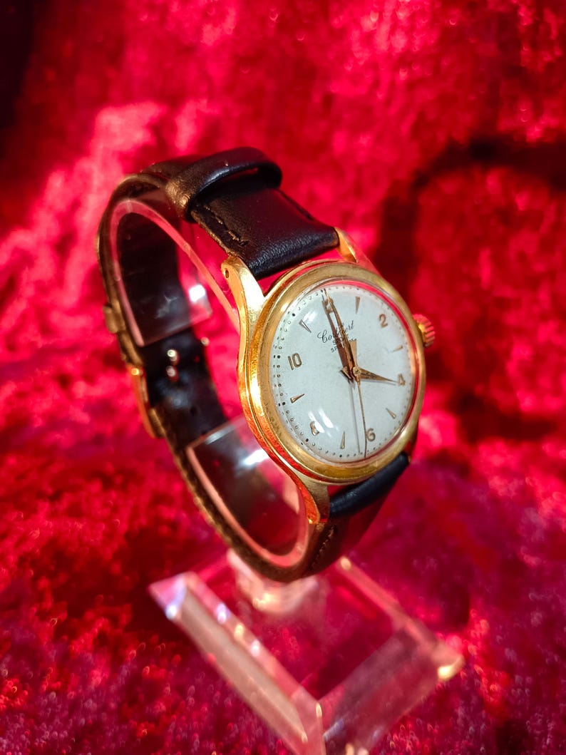 May include: A vintage gold-tone wristwatch with a white face and black leather band. The watch features gold hands and hour markers, with the numbers 2, 6, and 10 clearly visible. The watch is displayed on a clear stand against a red background.