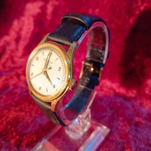May include: A vintage gold-toned wristwatch with a white face and black leather band. The watch face displays the brand name "Certus" and features gold hands and hour markers. The watch is displayed on a clear stand against a red background.