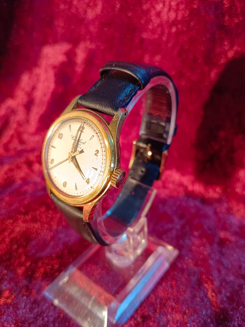 May include: A vintage gold-toned wristwatch with a white face and black leather band. The watch face displays the brand name "Certus" and features gold hands and hour markers. The watch is displayed on a clear stand against a red background.