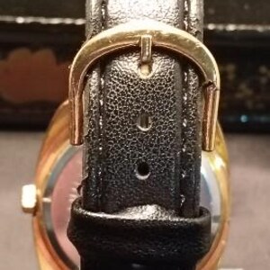 May include: Close-up of a gold-toned wristwatch with a black leather band. The band features a gold-colored buckle. The watch face is visible, with a gold-colored bezel and a light-colored dial.