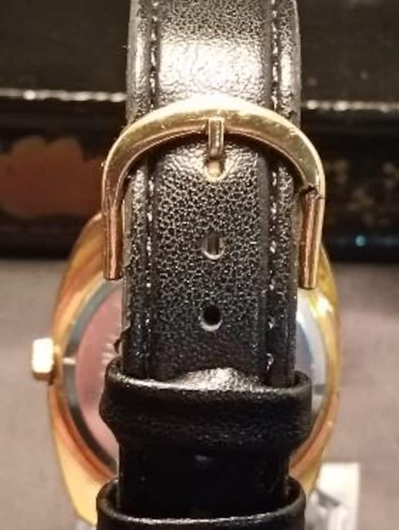 May include: Close-up of a gold-toned wristwatch with a black leather band. The band features a gold-colored buckle. The watch face is visible, with a gold-colored bezel and a light-colored dial.